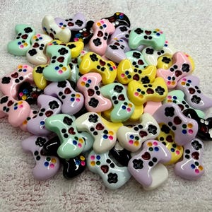 May include: A collection of colorful, miniature game controller charms in various pastel shades, including pink, yellow, green, and purple. Each charm features a black button layout and colorful accent buttons. The charms are scattered on a white, textured surface.