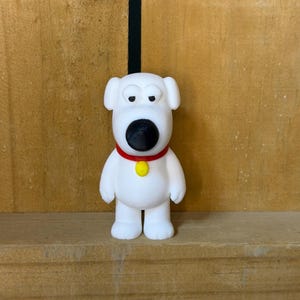 May include: A white anthropomorphic dog figurine with a large black nose, drooping eyes, and floppy ears. The figure wears a bright red collar featuring a small, circular yellow tag centered on the chest. The character stands upright with a smooth, matte finish.