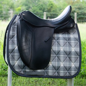 May include: A black leather saddle with a gray and white plaid saddle pad. The saddle is sitting on a white wooden stand.
