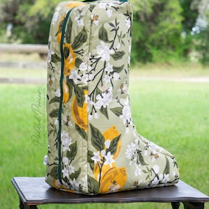May include: A tall, padded equine boot cover featuring a botanical design of lemons, white flowers, and bees on a pale green backdrop. It has dark green trim and a handle. The words "Padded Ponies" are printed on the side.