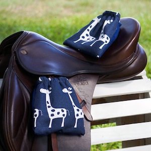 May include: A brown leather horse saddle with two blue and white fabric bags with a giraffe print. The bags are attached to the saddle with straps. The saddle is sitting on a white wooden bench.