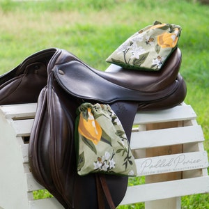 May include: Two saddle bags with floral patterns, one with lemons and the other with white flowers and bees, are placed on a brown leather saddle. The saddle is resting on a white wooden saddle stand. The text "Padded Ponies Custom Equine Accoutrements" is printed on the stand.