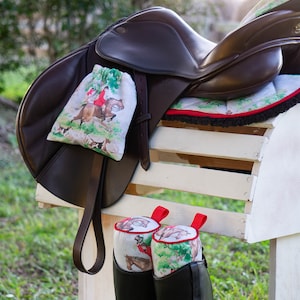 May include: A brown leather horse saddle with a white and green patterned saddle pad and a matching saddle bag. Two black riding boots with white and green patterned boot covers are hanging on a white wooden rack.