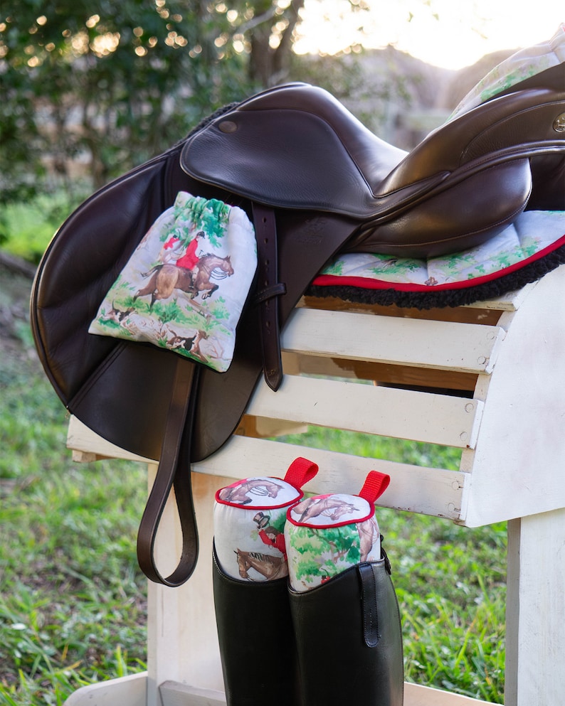 May include: A brown leather horse saddle with a white and green patterned saddle pad and a matching saddle bag. Two black riding boots with white and green patterned boot covers are hanging on a white wooden rack.