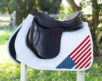 May include: A white saddle pad with a red, white, and blue American flag design. The saddle pad is on a saddle stand with a brown leather saddle on top.
