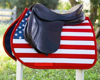 May include: A red, white, and blue saddle pad with a brown leather saddle on top. The saddle pad features a quilted design and the American flag.