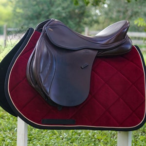 May include: A brown leather saddle with a burgundy velvet saddle pad. The saddle pad has a diamond quilted pattern and a white border.