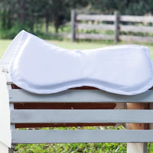 May include: A white saddle pad rests on a white wooden crate. The pad has a curved shape and is made of a smooth, padded material. The crate has horizontal slats and is set outdoors with a blurred green background.