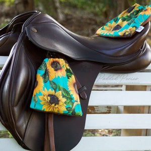 May include: A brown leather horse saddle with two turquoise and yellow sunflower print saddle bags. The saddle is sitting on a white wooden crate. The text "Padded Ponies Custom Equestrian Accoutrements" is visible on the saddle.