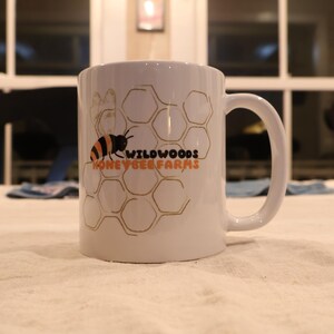 May include: White ceramic mug with a black and orange bee graphic and the words "Wildwoods Honey & Bee Farms" in orange and black. The mug features a honeycomb design and a large handle.