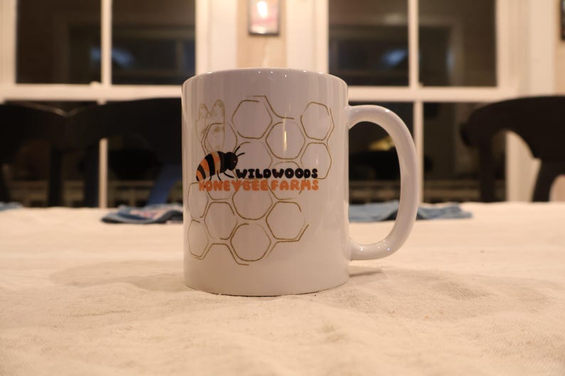 May include: White ceramic mug with a black and orange bee graphic and the words "Wildwoods Honey & Bee Farms" in orange and black. The mug features a honeycomb design and a large handle.