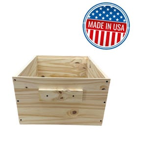 May include: A natural wood box with a rectangular shape. The box has a handle on the side and is made of light-colored wood. The words "MADE IN USA" are printed on a circular emblem.