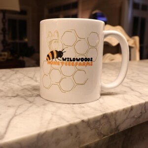 May include: White ceramic mug featuring a bee and honeycomb design with the text "Wildwoods Honeybee Farms" in black and orange. The mug has a curved handle and is sitting on a marble surface.