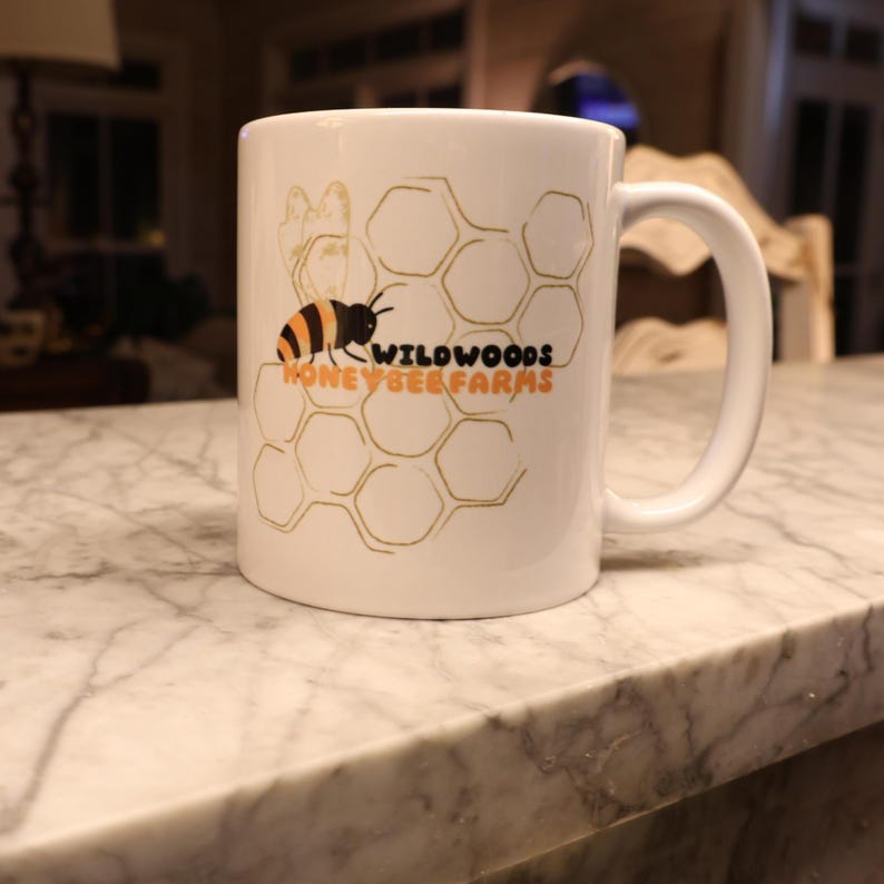 May include: White ceramic mug featuring a bee and honeycomb design with the text "Wildwoods Honeybee Farms" in black and orange. The mug has a curved handle and is sitting on a marble surface.