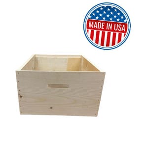 May include: A rectangular light-colored wooden crate with a horizontal cutout handle on the front panel. Above the crate is a circular emblem featuring the American flag design and the text MADE IN USA in bold white capital letters.