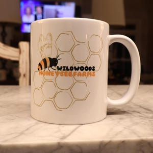 May include: White ceramic mug featuring a bee and honeycomb design with the text "WILDWOODS HONEY BEE FARMS" in orange and black. The mug has a curved handle and is set on a marble surface.