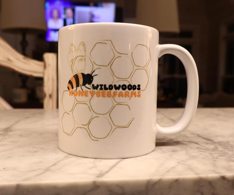 May include: White ceramic mug featuring a bee and honeycomb design with the text "WILDWOODS HONEY BEE FARMS" in orange and black. The mug has a curved handle and is set on a marble surface.