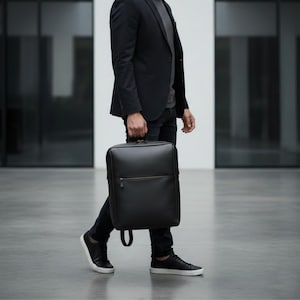 Puede incluir: Una mochila de cuero negro con forma rectangular y asa superior. La mochila tiene un bolsillo frontal con cremallera y un diseño minimalista. La persona lleva una blazer negra, pantalones negros y zapatillas negras.