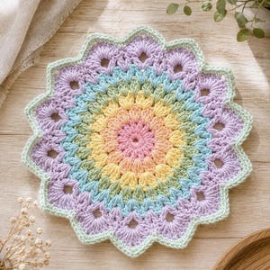 May include: A circular crochet doily featuring a radial pattern of pastel colors including pink, yellow, light blue, and lavender, finished with a scalloped light green border. The design consists of concentric rings of textured stitches.