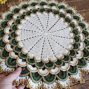 Puede incluir: Un tapete redondo de ganchillo con un patrón circular central blanco rodeado por capas concéntricas de bordes festoneados en verde oscuro y blanco, acabado con un borde de hilo dorado metálico en el contorno exterior y entre las capas de color.