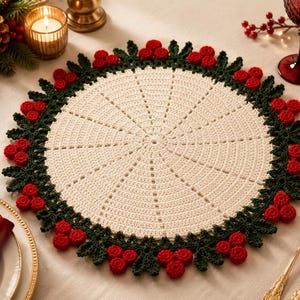 May include: A round, cream-coloured crocheted doily featuring a decorative border of dark green crocheted leaves and clusters of small, bright red crocheted berries arranged in a circular pattern around the outer edge of the textile piece.
