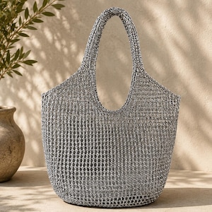 May include: A silver metallic tote bag featuring an open-weave crochet construction with a rounded base and two integrated handles. The bag has a structured yet flexible appearance with a visible grid pattern throughout the entire body.