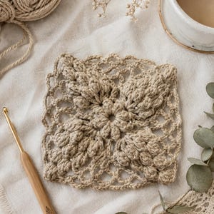 May include: A square crochet motif in a light beige yarn featuring a central flower design surrounded by four textured, raised leaf-like patterns and an openwork border. A wooden crochet hook with a gold metal tip is positioned to the left.