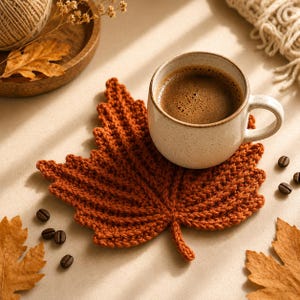 May include: A rust-colored crochet coaster shaped like a maple leaf, supporting a speckled cream-colored ceramic mug filled with dark coffee. The coaster features detailed ribbed stitching that mimics the veins of a leaf, providing a textured surface.