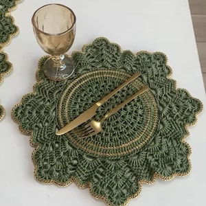 May include: A round, sage green crocheted placemat with a scalloped edge and metallic gold thread trim. A gold-toned knife and fork are placed in the center of the placemat, accompanied by a clear, stemmed glass positioned on the upper left side.