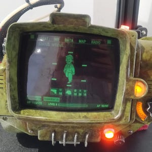 Pode incluir: Uma réplica personalizada, verde-oliva e castanho, do Pip-Boy 3000 da série de videojogos Fallout. O ecrã exibe um gráfico de personagem verde e dados do jogo. O dispositivo tem vários botões e botões, e uma pequena luz vermelha.