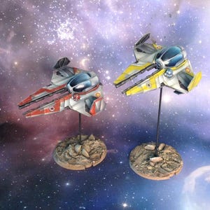 May include: Two miniature spacecraft models, one red and white, the other yellow and white, are displayed on stands against a cosmic background. The models have detailed designs and are mounted on textured, rocky bases.