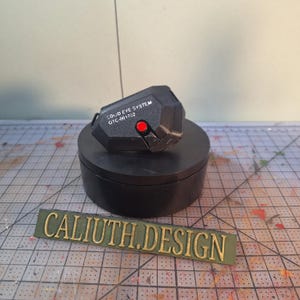 Op de afbeelding: Een zwart, geometrisch apparaat met een rode knop en de tekst "SOLID EYE SYSTEM OTC-001702" wordt weergegeven op een zwarte, cirkelvormige standaard. De basis van de standaard is ook zwart. Een groen rechthoekig bord met gouden letters met de tekst "CALIUTH.DESIGN" staat op de voorgrond.