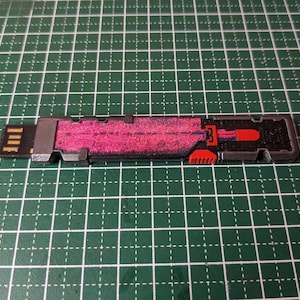 Könnte beinhalten: Rechteckiger USB-Stick mit schwarzem USB-Anschluss und einem Gehäuse mit rosa und rotem Design. Der Stick hat einen schwarz-grauen Rahmen mit roten Akzenten, vor einem grünen Gitterhintergrund.