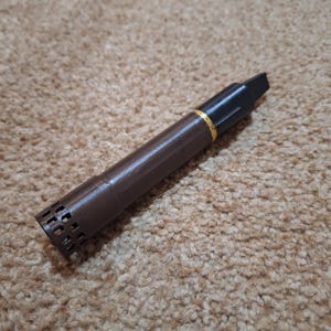 Op de afbeelding: Een bruin en zwart penvormig object met een gouden band. De pen heeft een getextureerde bruine behuizing en een zwarte punt. Het uiteinde van de pen heeft een reeks kleine rechthoekige openingen.