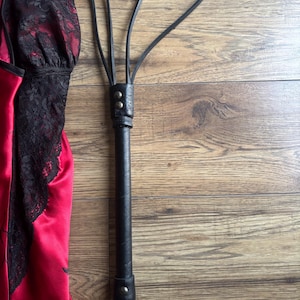 Puede incluir: Un flagelador de cuero negro con múltiples caídas y un asa en forma de lazo. El flagelador está junto a una prenda de satén rojo con ribete de encaje negro. Los artículos están sobre una superficie de madera.