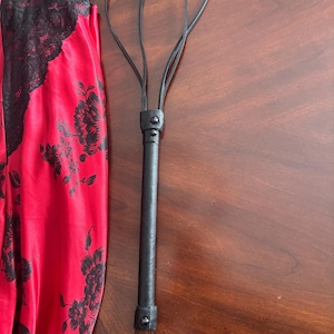 May include: A black leather flogger with a looped handle and a long, textured handle. Next to it is a red satin garment with black floral patterns and black lace trim. The items are on a wooden surface.