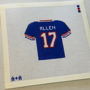 May include: A needlepoint canvas featuring a blue sports jersey design with the name ALLEN and the number 17 in white with red outlines. The jersey has red and white striped sleeve cuffs. The text A+A is in the bottom left corner.