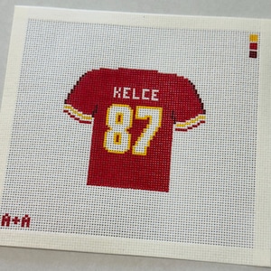 May include: A square needlepoint canvas featuring a red football jersey design with the name KELCE and the number 87 in white with yellow outlines. The design includes small color swatches in the top right and the text A+A in the bottom left corner.