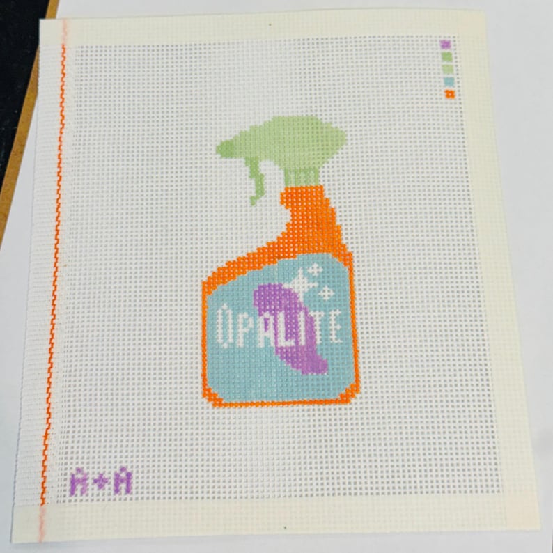 May include: Needlepoint canvas with a spray bottle design. The bottle is orange with a light green spray nozzle and a light blue label with the word "OPALITE" and a purple design. The canvas has a white background.