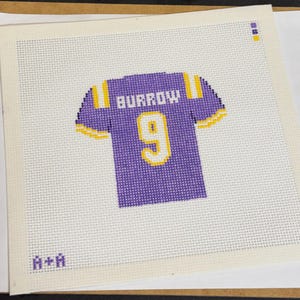 Op de afbeelding: Een kruissteekontwerp van een paars en goud voetbalshirt met de naam "BURROW" en het nummer "9". Het shirt staat op een witte achtergrond met een rasterpatroon. De initialen "A+A" staan in de linkerbenedenhoek.