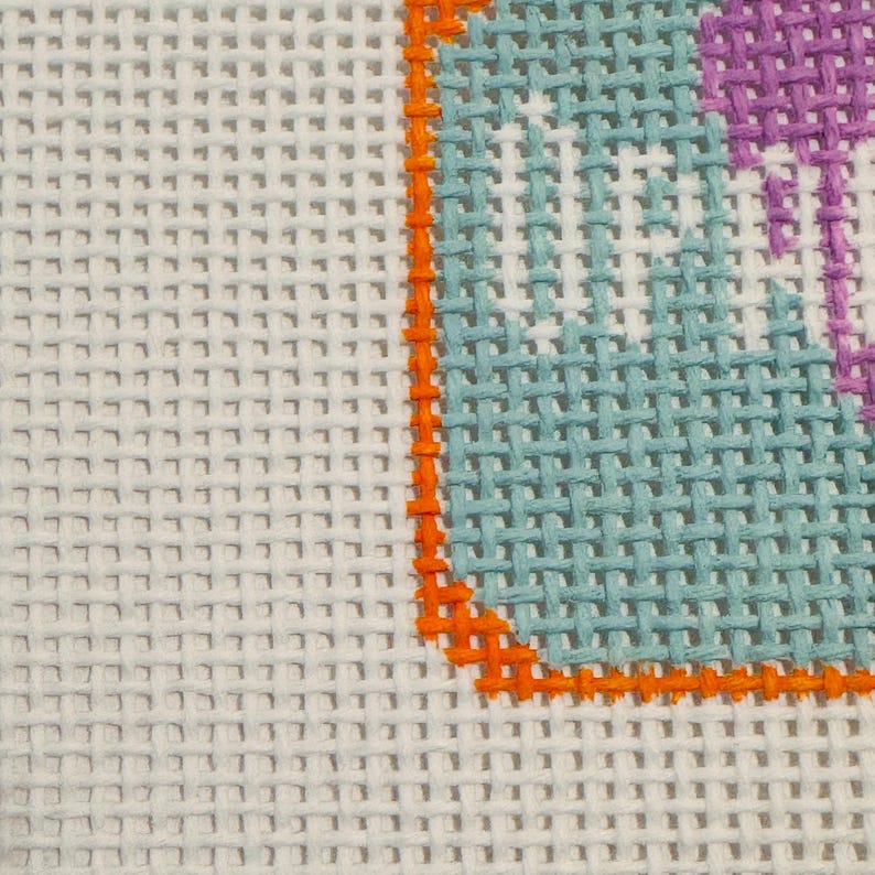 May include: Close-up of a needlepoint canvas with a white background. The design features a section of a light blue shape outlined in orange, with a purple section. The canvas has a grid-like texture.