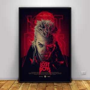 May include: A framed movie poster for "The Lost Boys" featuring a portrait of a man with a split face, half human and half vampire, against a red background. The title is in red, with the director's name at the top.