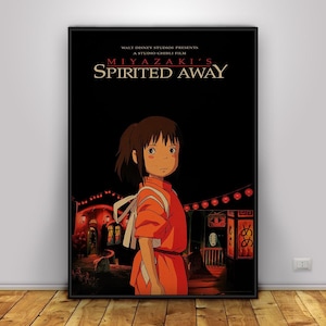 May include: A framed poster for the animated film "Spirited Away" by Studio Ghibli. The poster features a young girl in an orange dress, with the film's title and credits in white text against a black background. The scene includes lanterns and buildings.