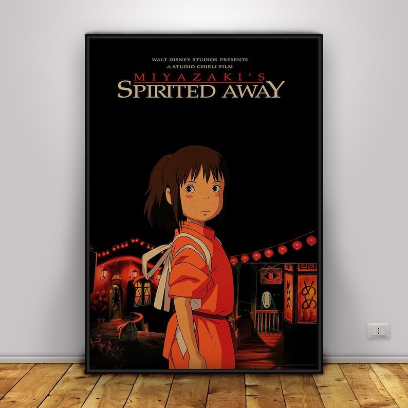 May include: A framed poster for the animated film "Spirited Away" by Studio Ghibli. The poster features a young girl in an orange dress, with the film's title and credits in white text against a black background. The scene includes lanterns and buildings.