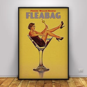 May include: A framed poster featuring a woman reclining in a martini glass, holding an olive on a toothpick. The poster has a yellow background with the title "FLEABAG" and the name "PHOEBE WALLER-BRIDGE" at the top. The woman is wearing a dark dress and black heels.