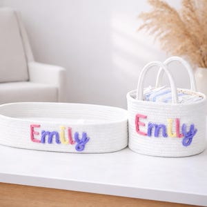 May include: Two white woven storage baskets. One is oval, the other round with handles. Both have the name "Emily" embroidered in colourful thread. The baskets are on a white surface.