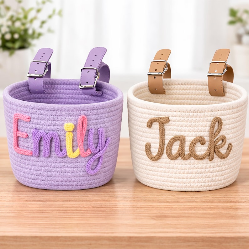 May include: Two woven rope baskets with leather straps and metal buckles. One basket is lavender with the name "Emily" in colorful yarn. The other is cream-colored with the name "Jack" in brown yarn. Both are sitting on a wooden surface.