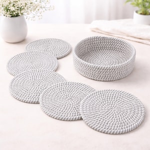 Könnte beinhalten: Ein Set aus sechs grau-weißen gewebten Untersetzern und einem passenden runden Korb. Die Untersetzer sind kreisförmig, mit einem Spiraldesign. Der Korb hat eine ähnliche gewebte Textur. Die Artikel deuten auf ein Wohnkulturthema hin.