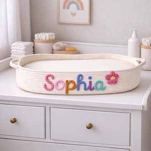 Può includere: Un cestino per il cambio del bambino color crema con il nome "Sophia" scritto con lettere di filo colorate. Un fiore rosa e giallo è accanto al nome. Il cestino è su un comò bianco con pomelli dorati.