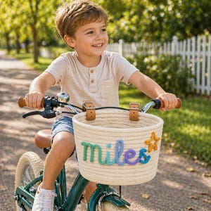 May include: A child riding a green bicycle with a woven basket. The basket is adorned with the name "Miles" in colorful yarn and a star. The bicycle has brown leather accents and a silver bell.